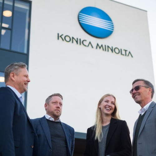 Several Konica Minolta employees in suits stand in a circle in front of an office building.